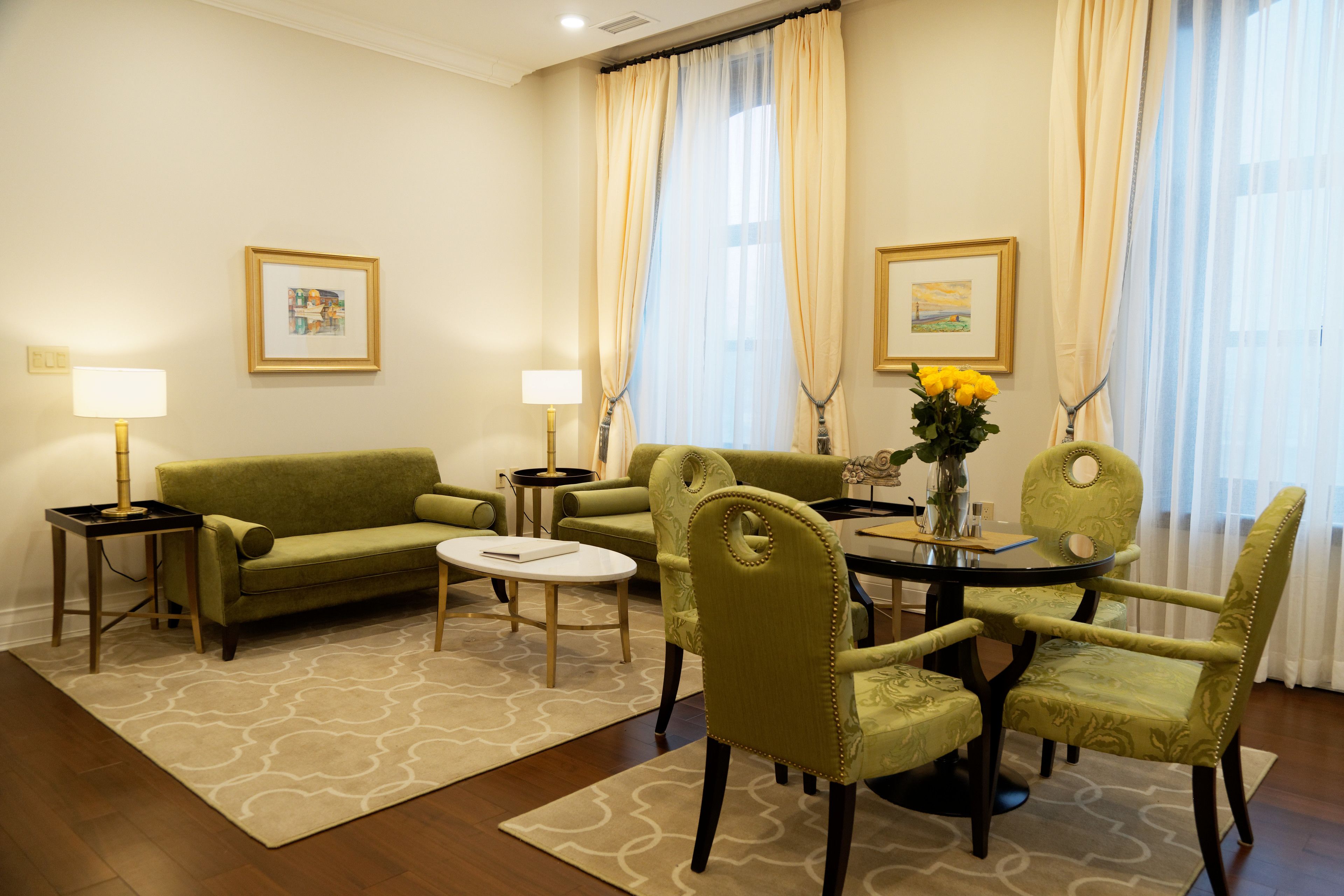 Habitación de hotel con sofá verde, dos sillones, una mesa de comedor con sillas, un jarrón con flores y dos cuadros en la pared.