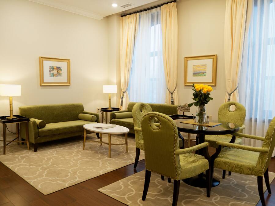 Habitación de hotel con sofá verde, dos sillones, una mesa de comedor con sillas, un jarrón con flores y dos cuadros en la pared.