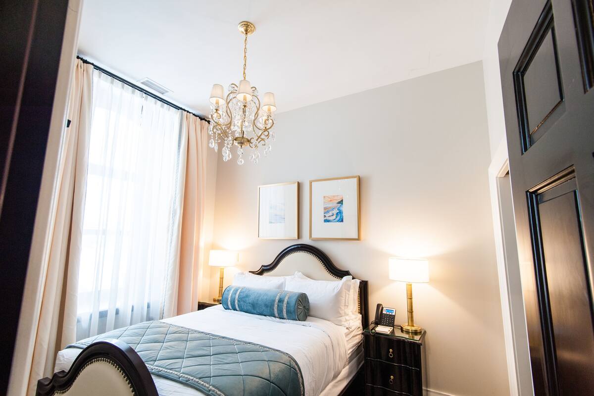 A hotel room with a bed, a chandelier, two framed pictures on the wall, and a nightstand with a lamp.