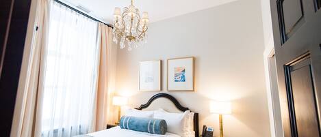 Habitación de hotel con una cama, una araña, dos cuadros enmarcados en la pared y una mesita de noche con lámpara.