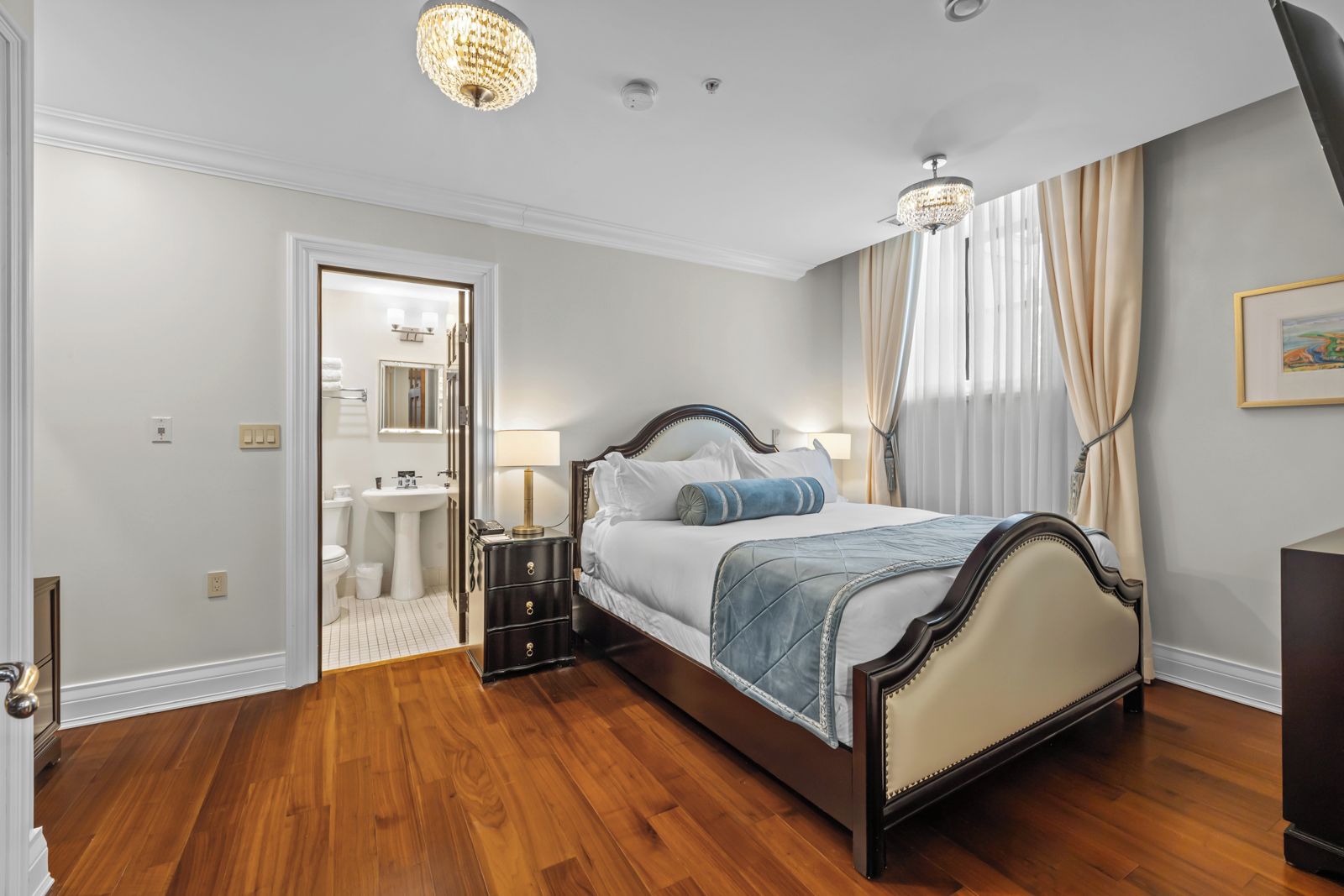 A bedroom with a wooden floor, a bed with a headboard, bedside tables, a nightstand, a chandelier, and a door leading to a bathroom.