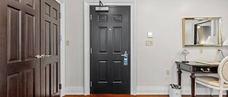 A hallway with a dark wooden door, a white wall, a small trash can, and a desk with a chair.