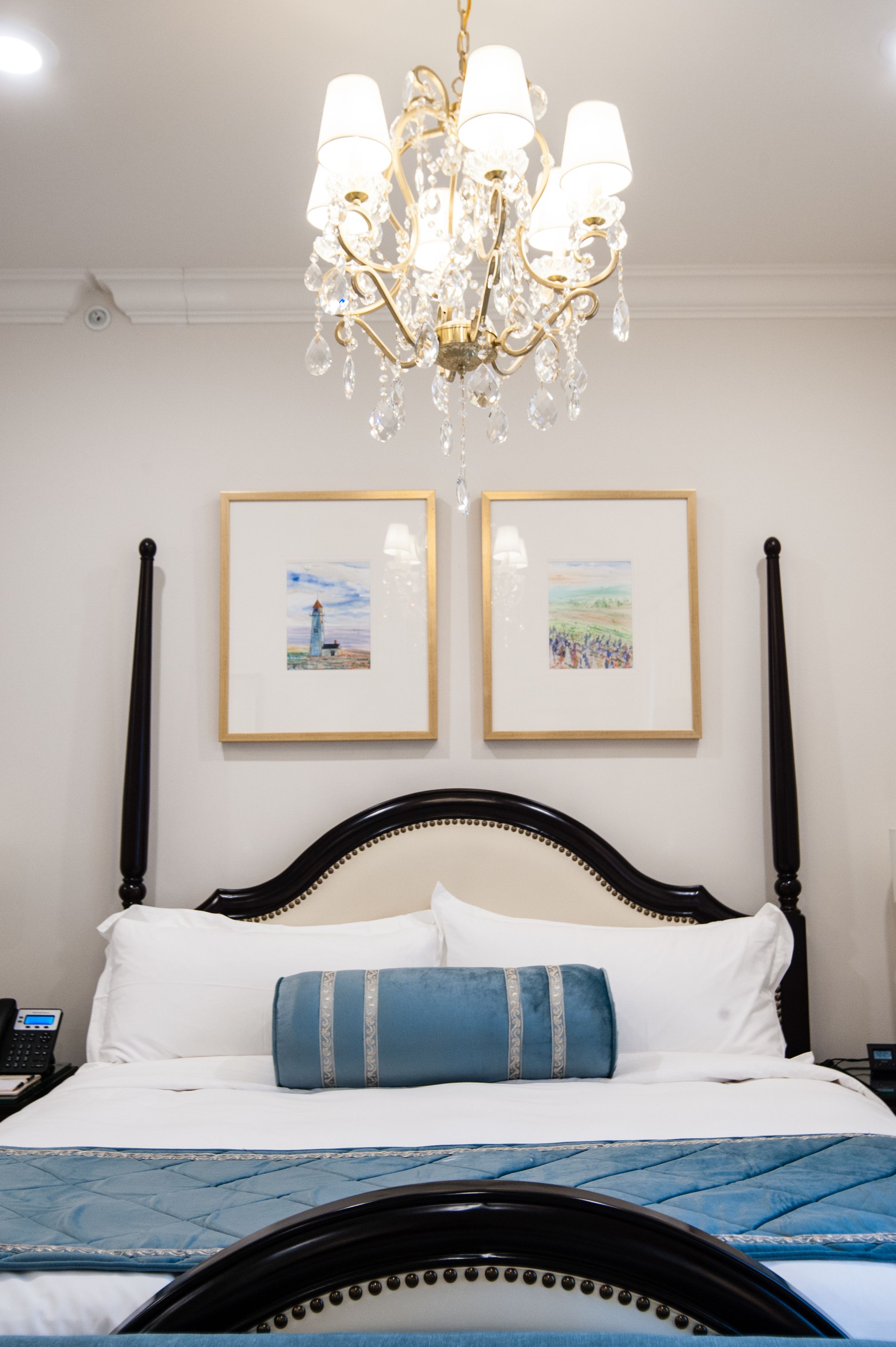 A neatly made bed with a blue quilt, a black headboard, and a chandelier above. There are two framed pictures on the wall.
