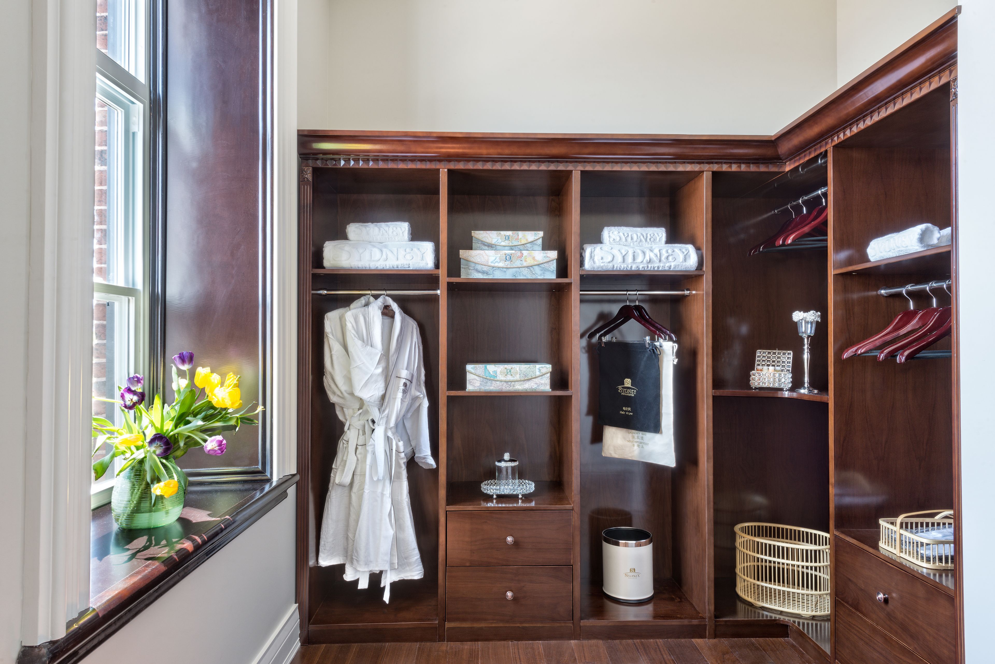 Un vestidor con estanterías de madera, una bata colgada y un jarrón con flores junto a la ventana.