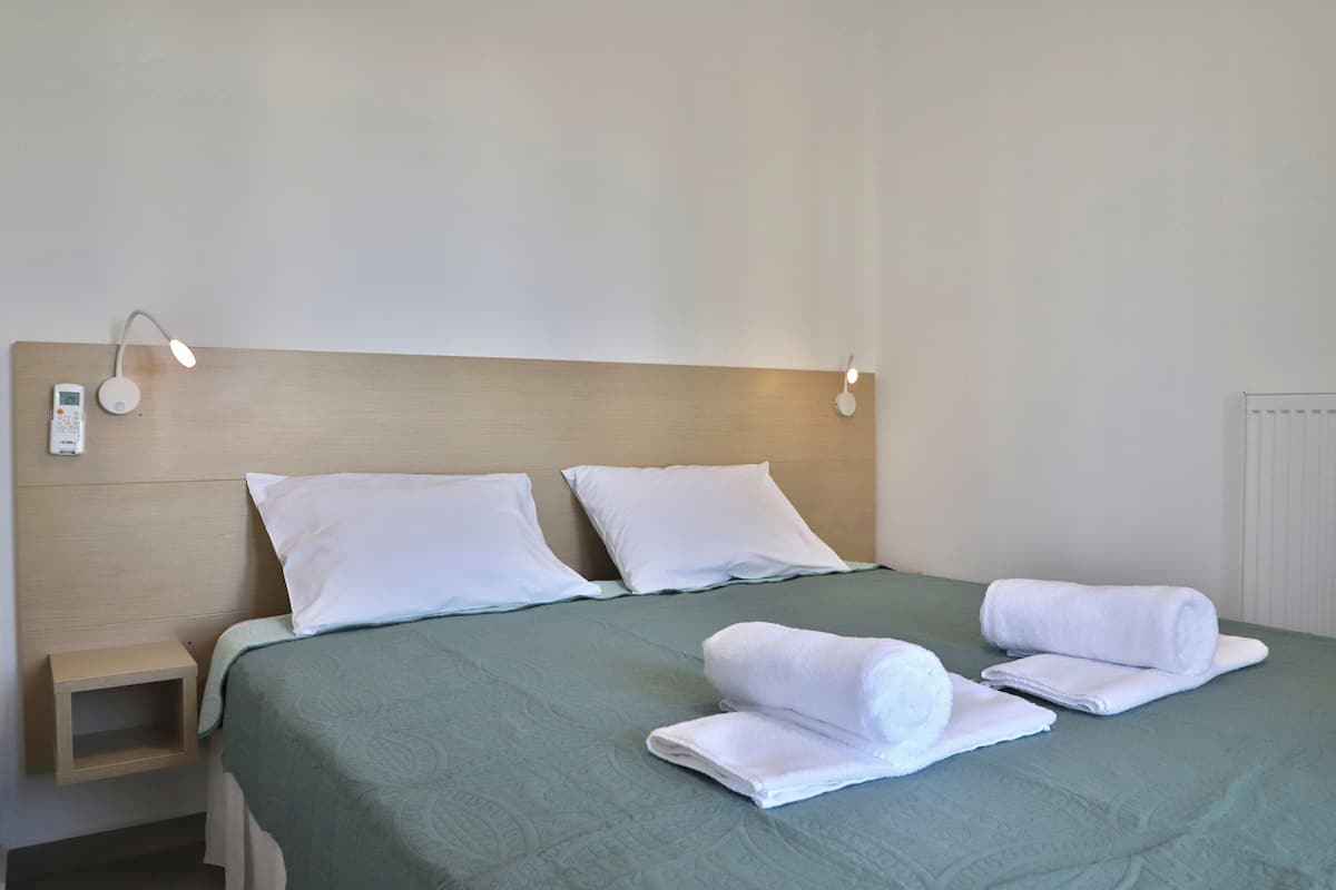 A neatly made bed with a green bedspread, two white pillows, and two white towels folded on the bed. There is a small wooden nightstand with a lamp beside the bed.