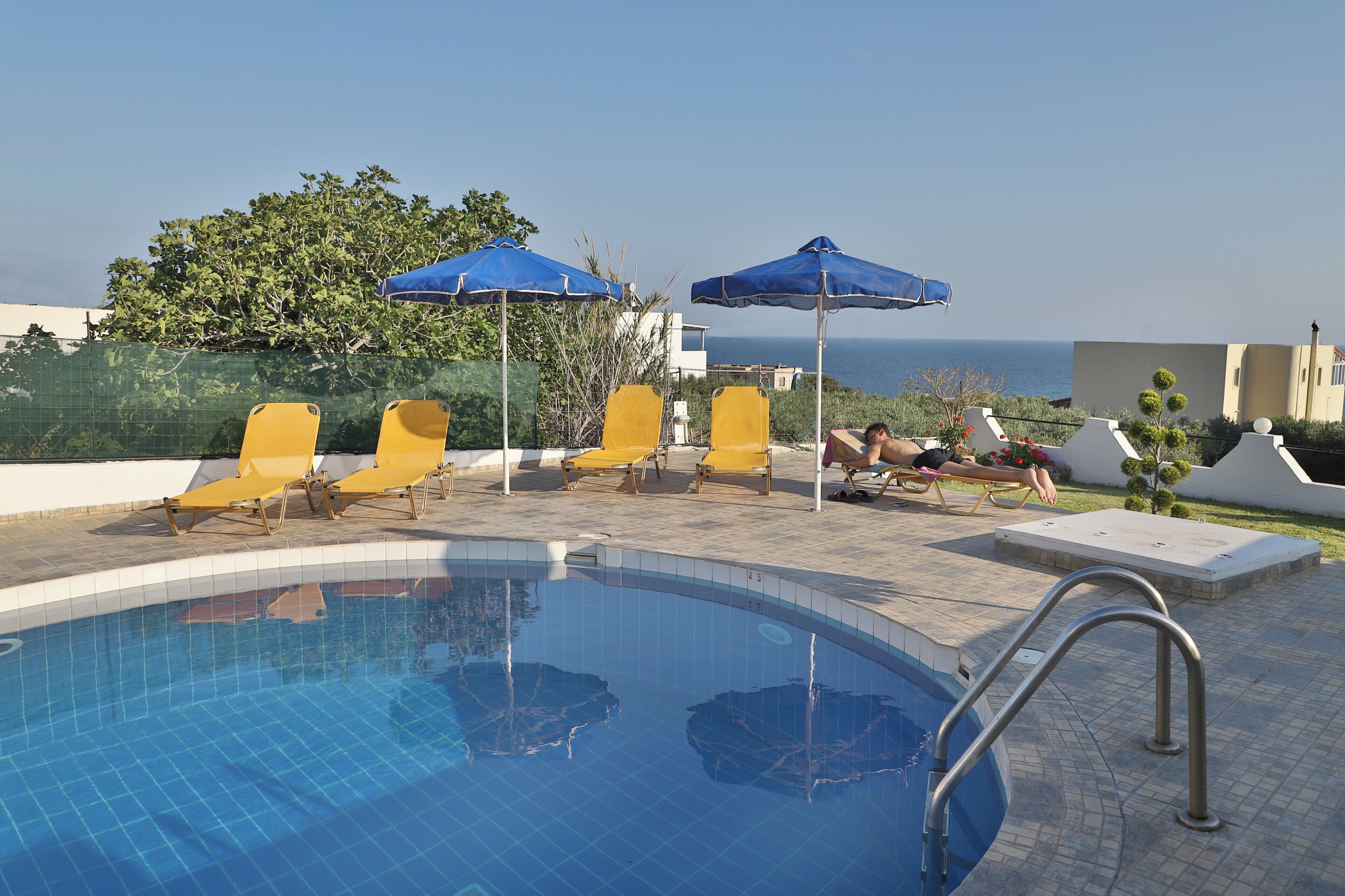 A swimming pool with yellow lounge chairs and blue umbrellas, surrounded by a tiled deck and a view of the sea.