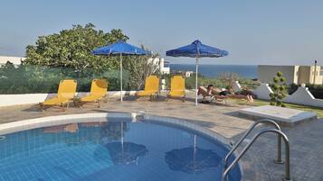 A swimming pool with yellow lounge chairs and blue umbrellas, surrounded by a tiled deck and a view of the sea.