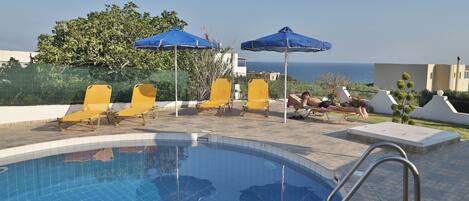 A swimming pool with yellow lounge chairs and blue umbrellas, surrounded by a tiled deck and a view of the sea.