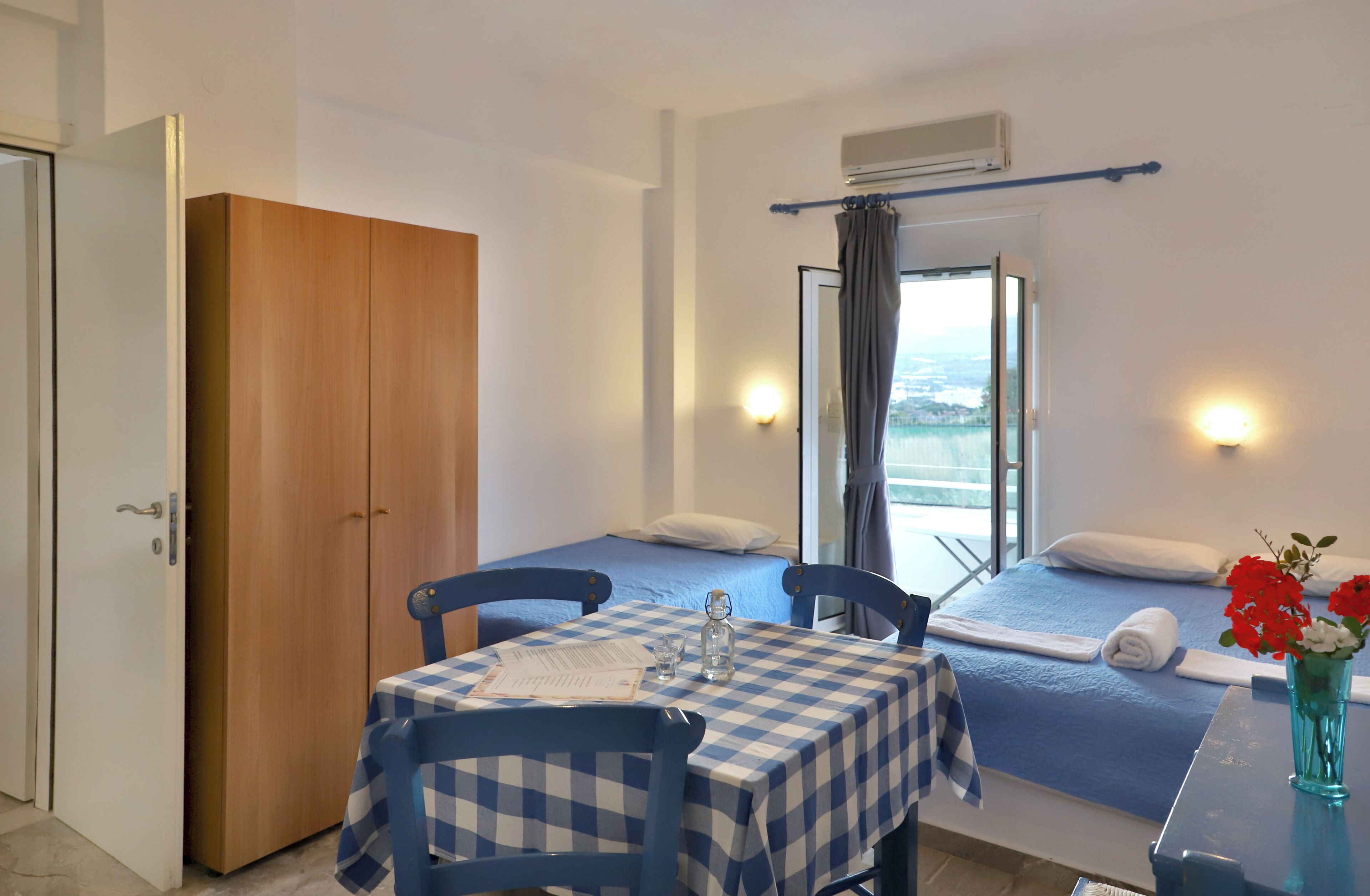 A room with a bed, a table with a checkered tablecloth, a chair, a wardrobe, and a window with curtains.