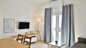 Een moderne, goed verlichte kamer met een houten eettafel, een klein wit bureau, een aan de muur bevestigde televisie, een lamp en grijze gordijnen voor een glazen schuifdeur.