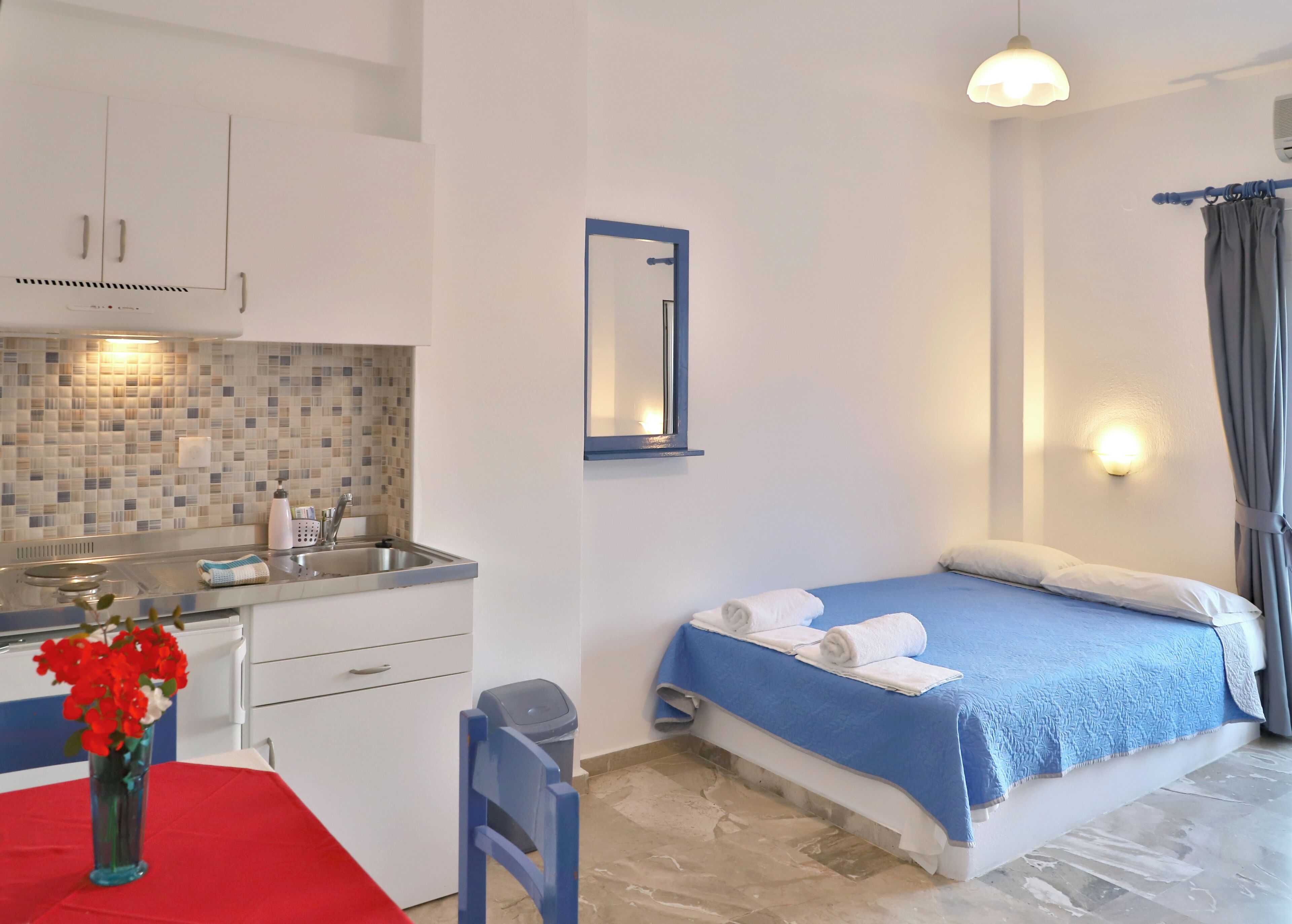 A compact hotel room with a kitchenette, a bed with blue bedding, a small table with a red tablecloth, and a vase with red flowers.