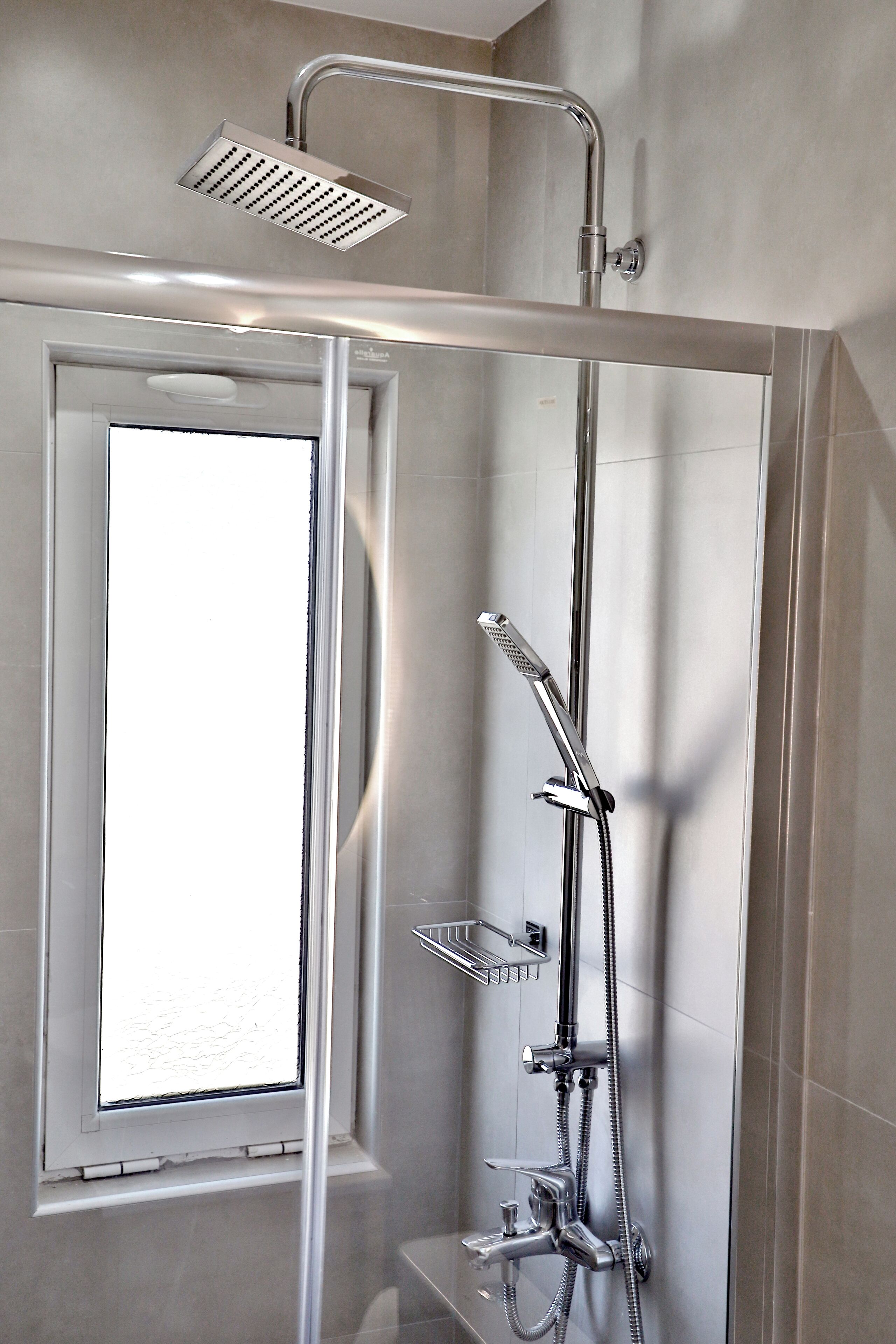 A modern shower enclosure with a glass door, a handheld showerhead, a wall-mounted faucet, and a window for natural light.