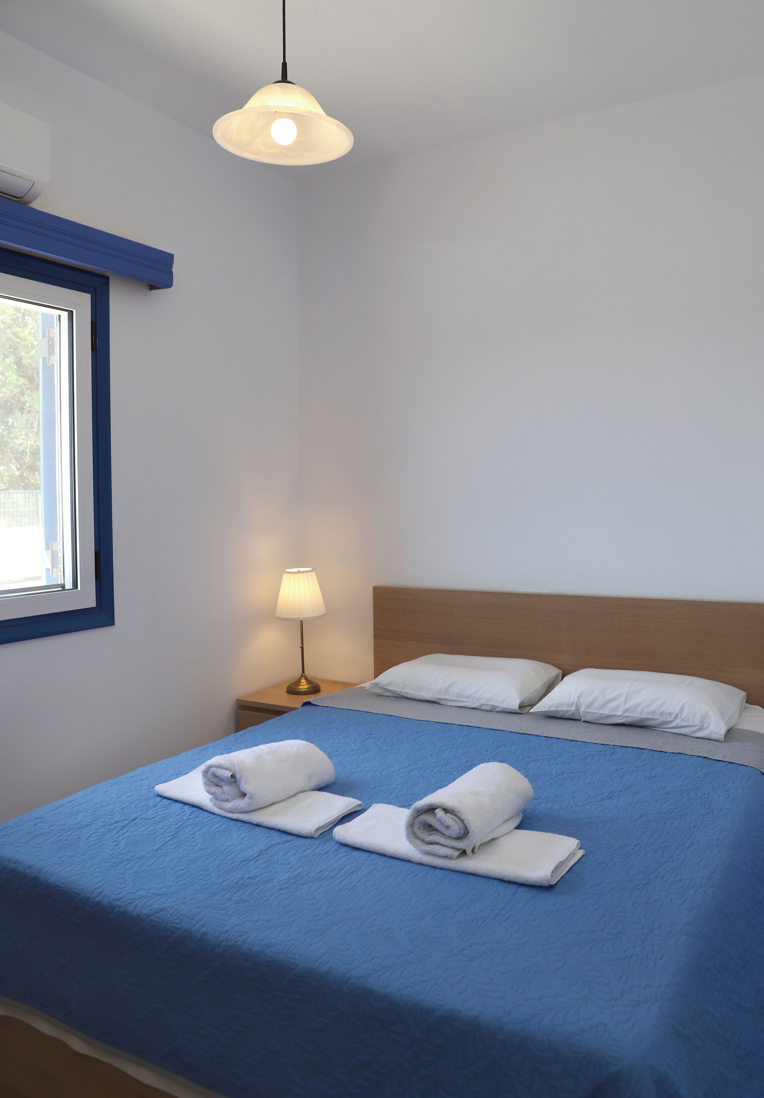 A neatly made bed with blue bedding and white pillows, a bedside lamp, and a window with blue trim.