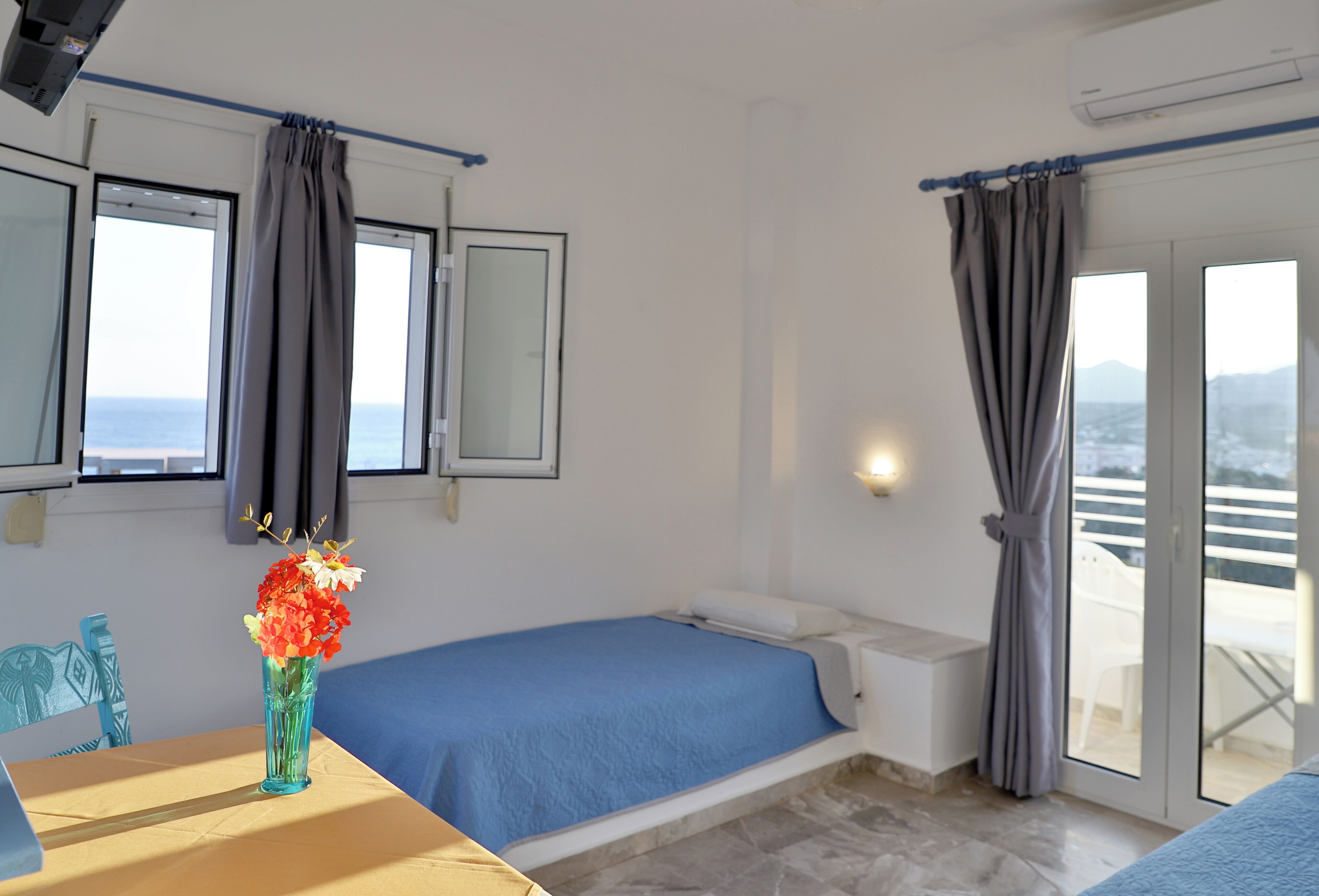 Una habitación con cama, una mesa con un jarrón de flores, una ventana con cortinas y vista al mar.