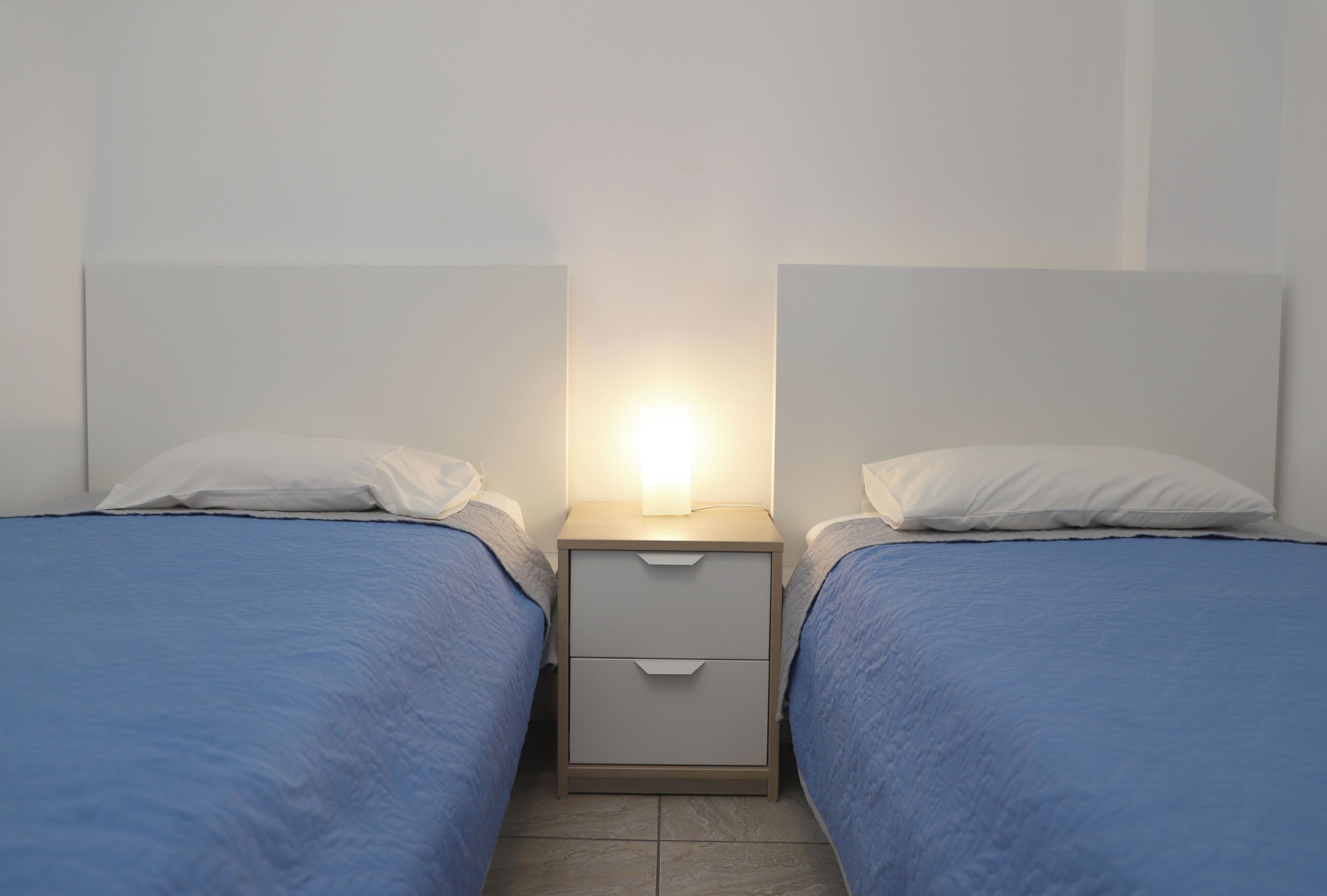 Habitación doble con dos camas, cada una con cabecero blanco y colcha azul. Hay una mesita de noche con lámpara entre las camas.