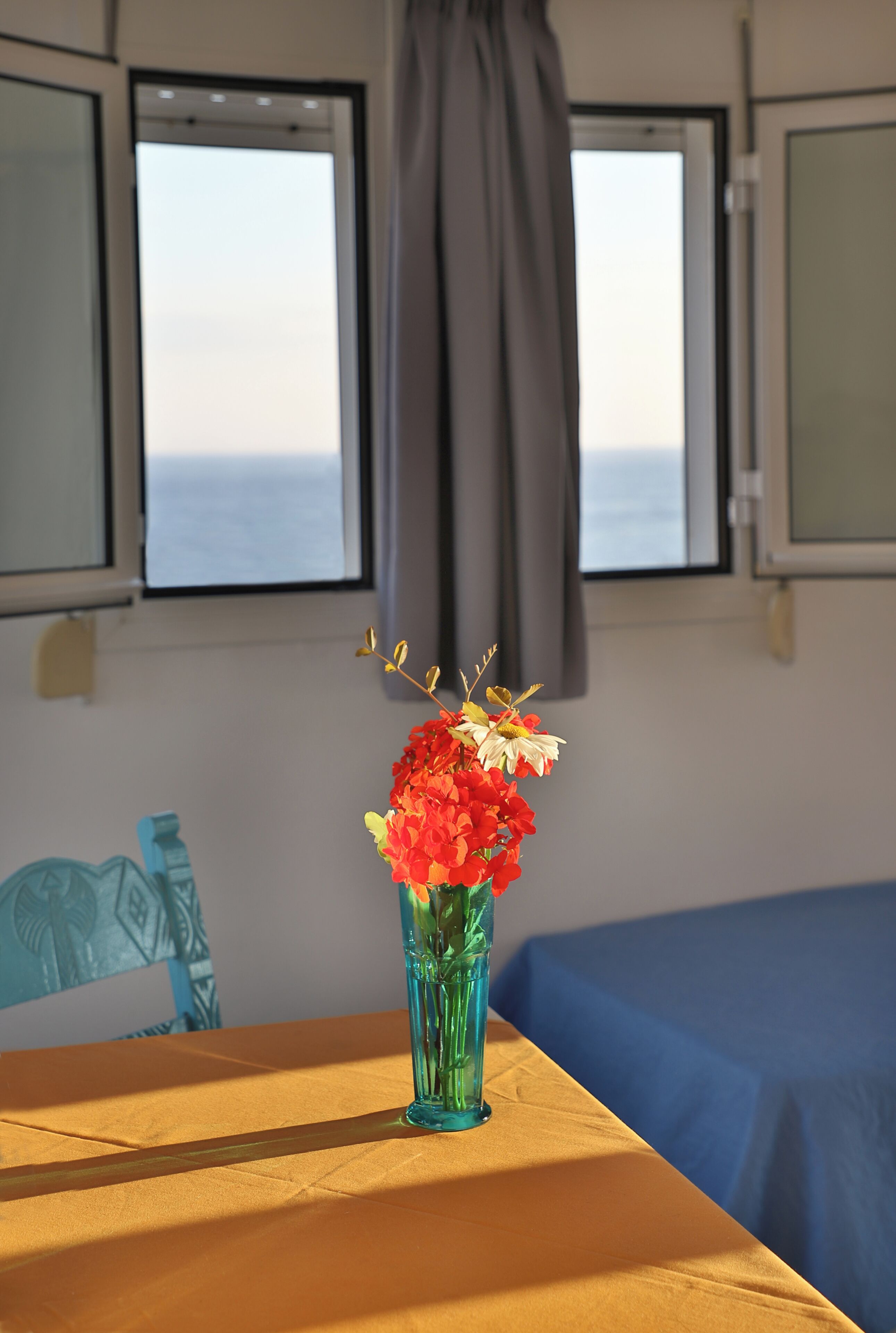 A room with a table, a vase with flowers, a window with curtains, and a bed with a blue cover.