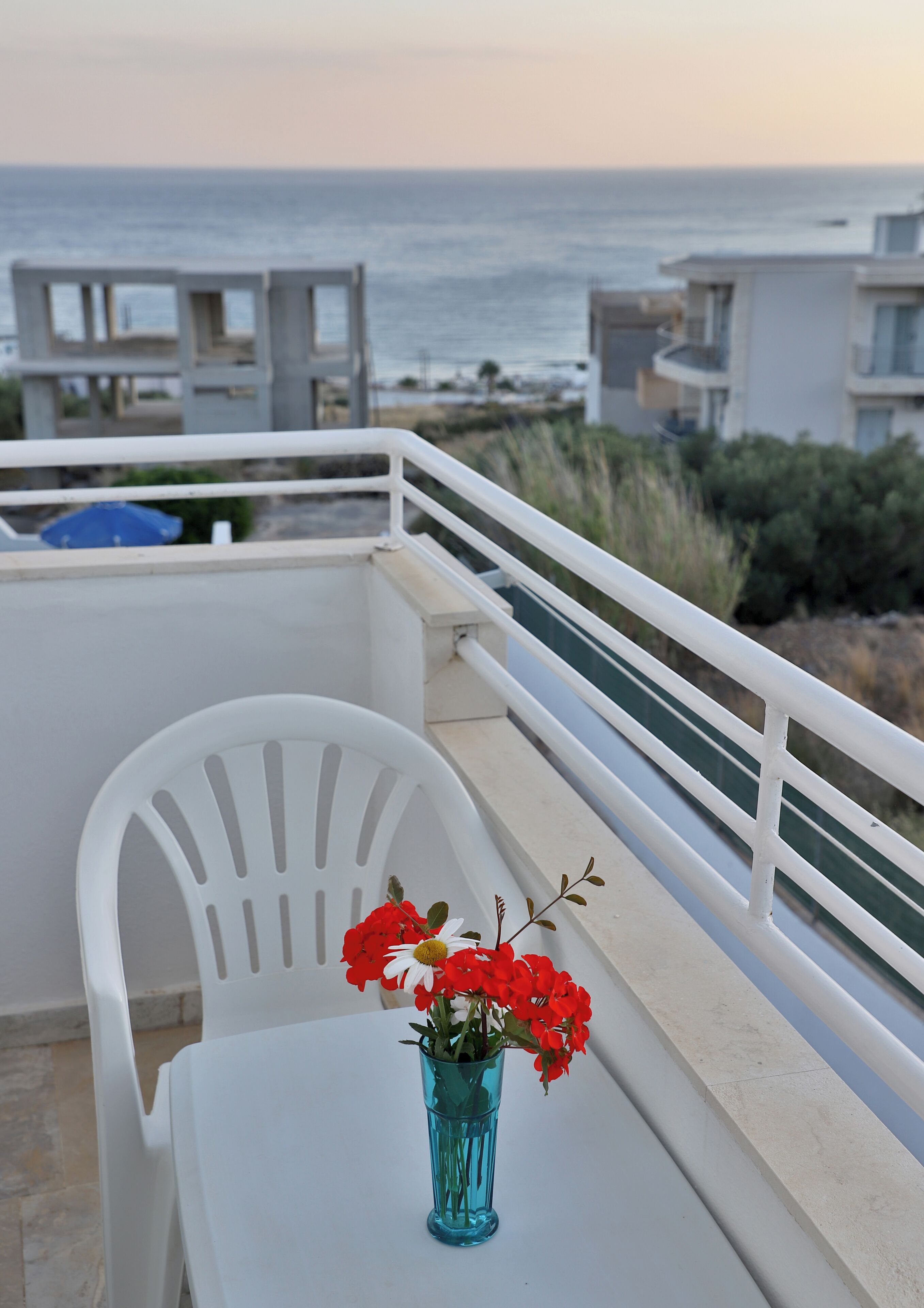 A balcony with a white chair, a small table, and a vase with red flowers. The background features a view of the sea and buildings.