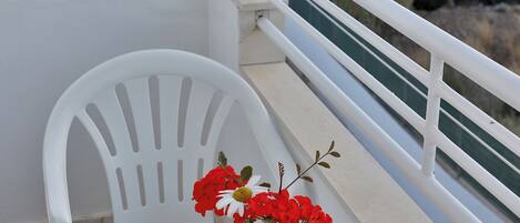 A balcony with a white chair, a small table, and a vase with red flowers. The background features a view of the sea and buildings.