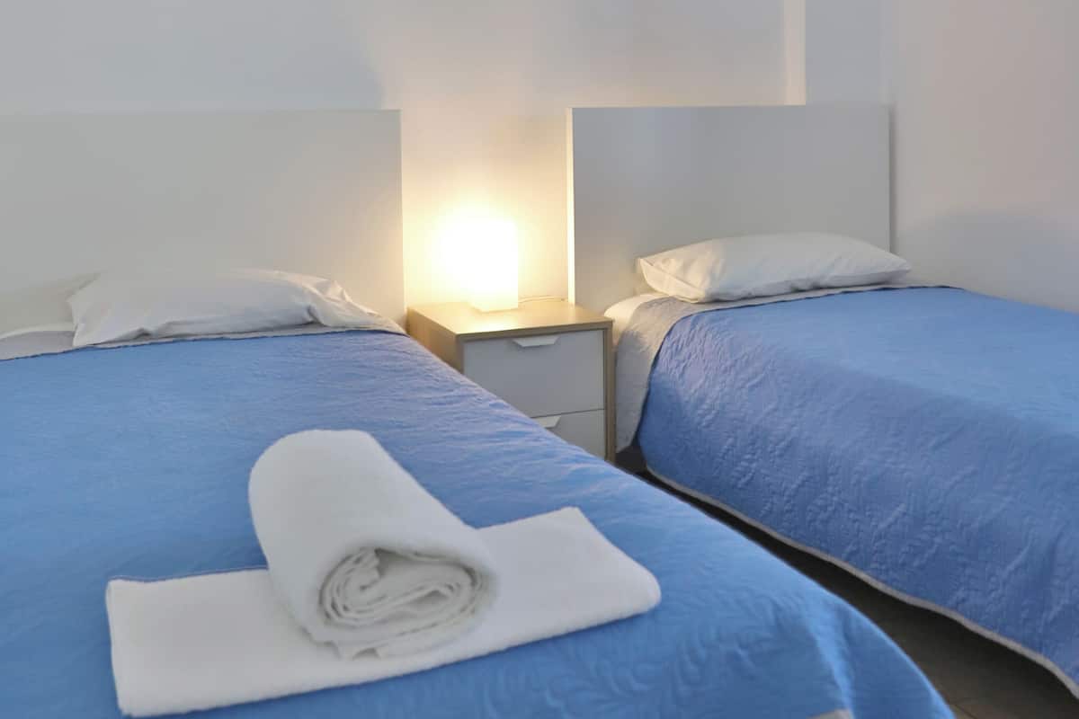 A hotel room with two beds, each with a blue bedspread and white pillows. There is a nightstand with a lamp between the beds.