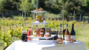 A table with a three-tiered stand holding pastries, jars of preserves, and bottles of wine, set against a vineyard backdrop.