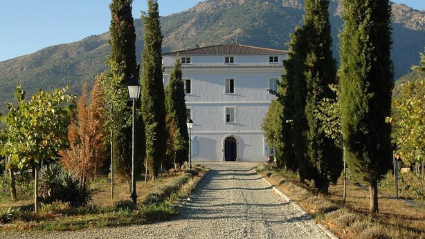A building with a central entrance, flanked by tall trees, set against a mountainous backdrop.