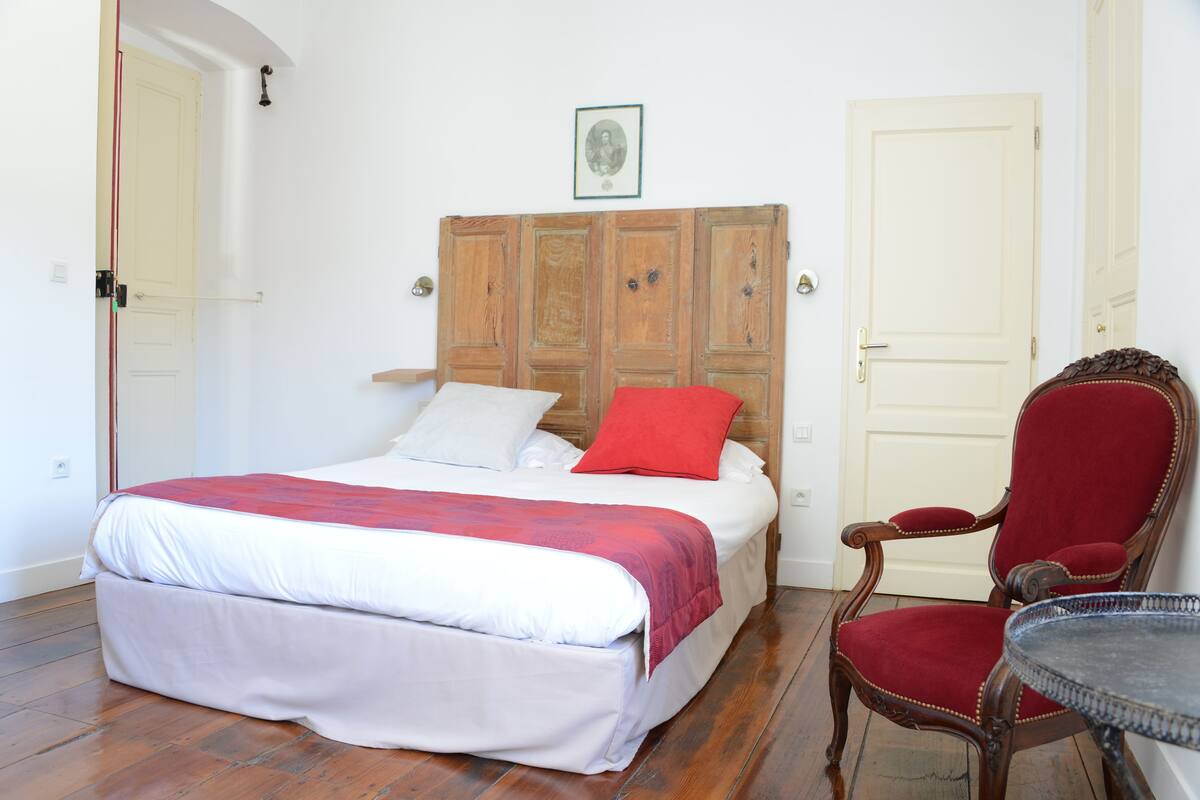 Een slaapkamer met een houten hoofdbord, een bed met witte en rode beddengoed, een rode fauteuil en een kleine ronde tafel.