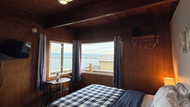 Ett mysigt rum med ett träpanelat tak, en säng med ett rutigt överkast, ett bord med en stol och en utsikt över havet genom fönstret.