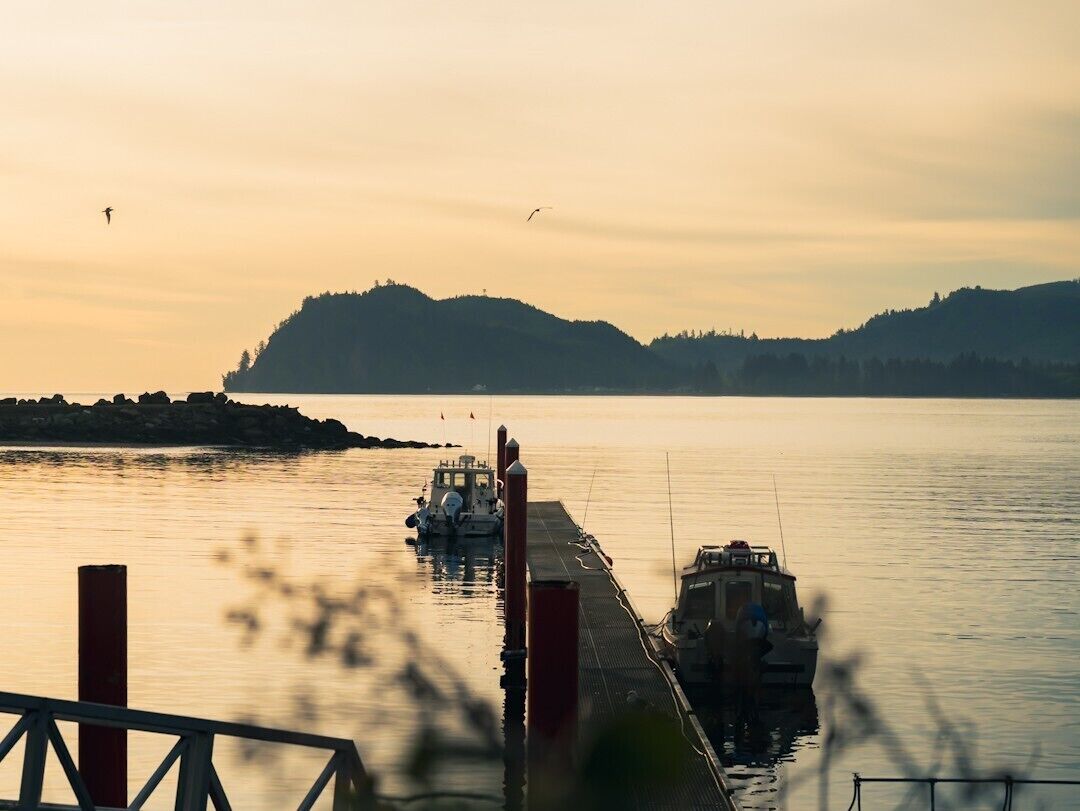 Una orilla tranquila con barcos amarrados, un muelle que se adentra en el agua y un paisaje montañoso al fondo.