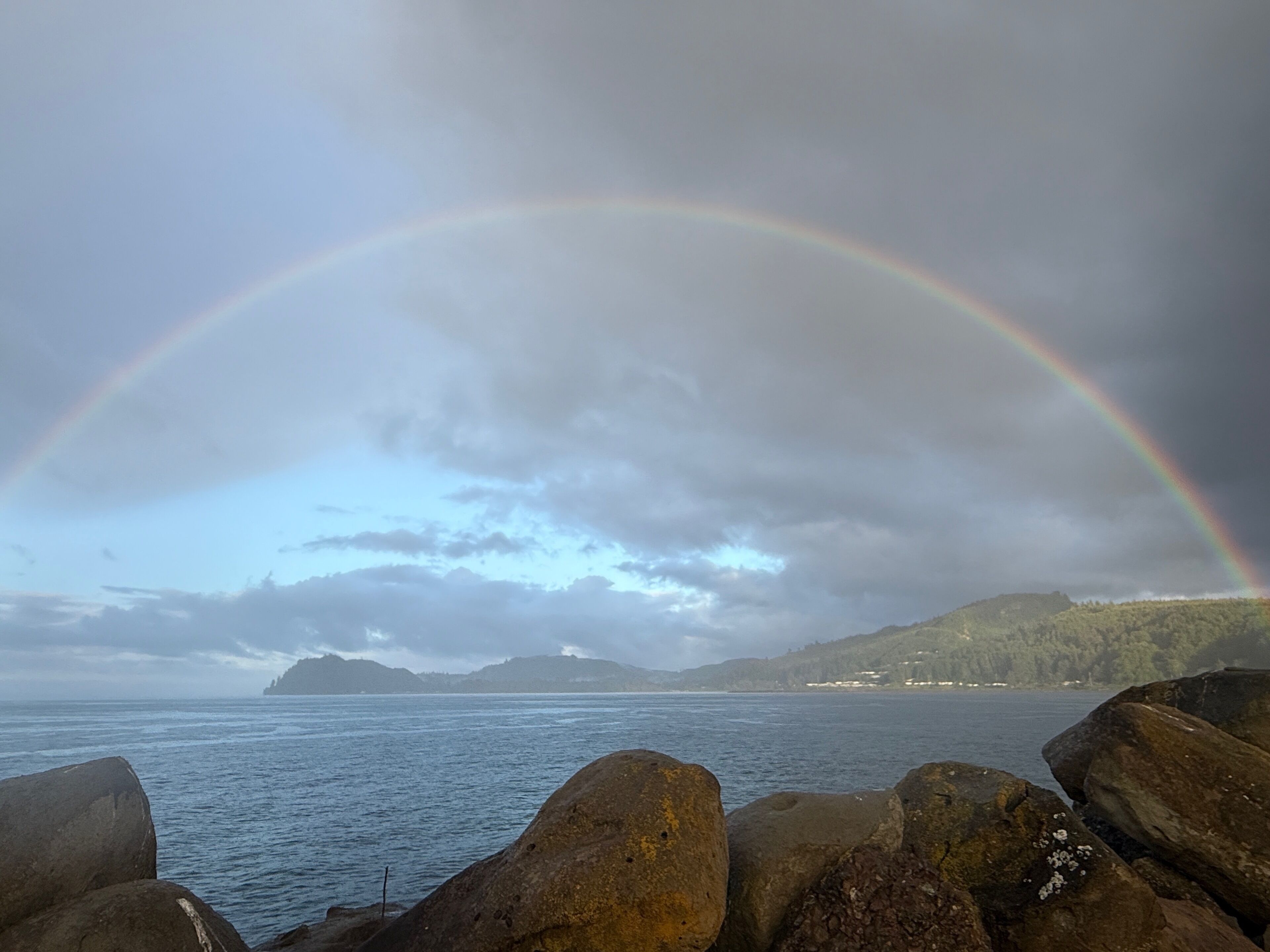 A coastal landscape with a rainbow over water, large rocks in the foreground, and a hilly background.