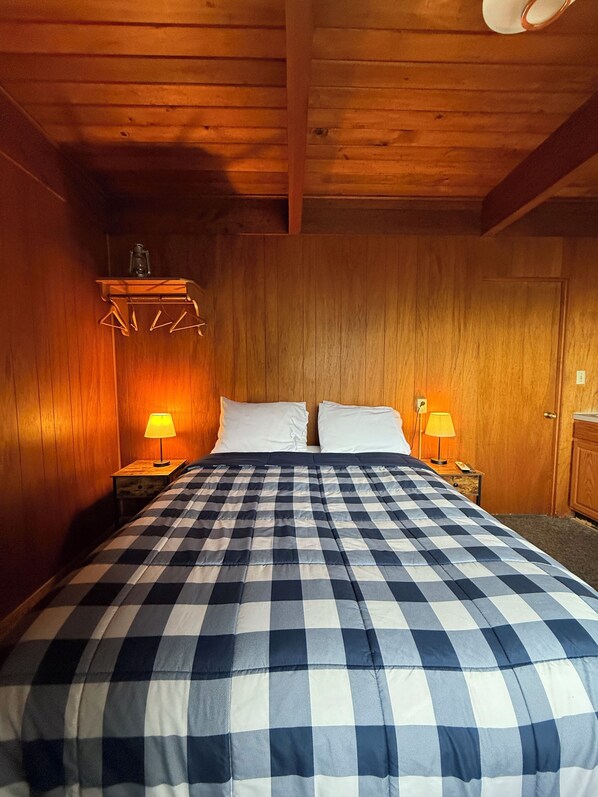 A room with wooden walls, a checkered bedspread, two bedside lamps, and a small shelf with a glass container.