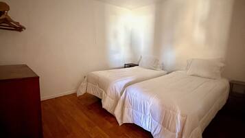 A room with two beds, a wooden floor, a ceiling light fixture, and a small table.
