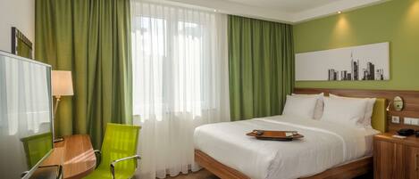 A hotel room with a bed, a desk with a chair, a TV, and a cityscape painting on the wall.