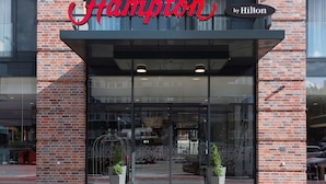 Entrada de un hotel Hampton by Hilton, con fachada de ladrillo, puertas de cristal grandes y un letrero rojo llamativo.