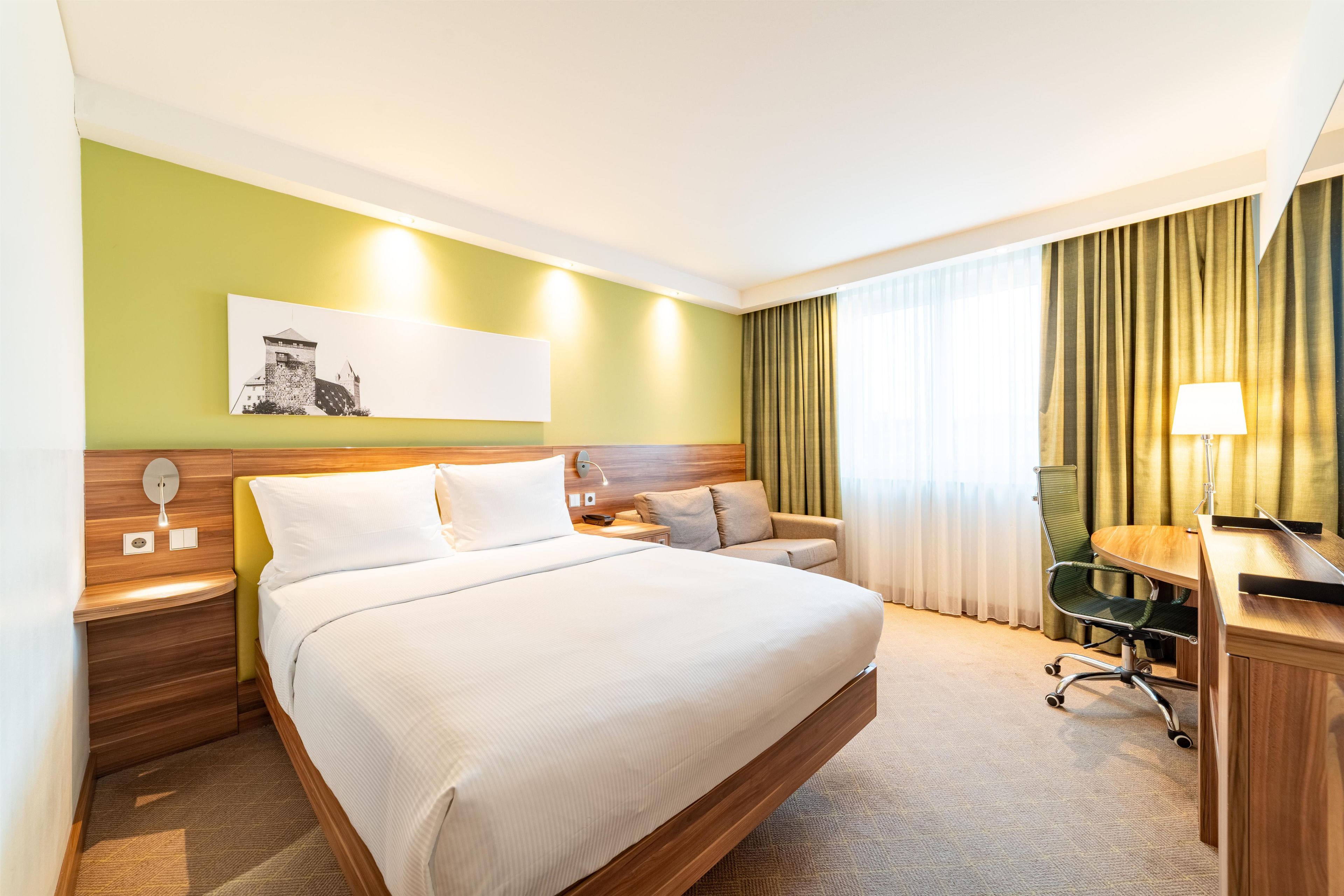 A hotel room with a large bed, a desk, a chair, a lamp, and a painting on the wall.