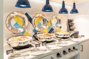 A buffet setup with labeled food stations including meatballs, baked beans, and grilled vegetables. - Hampton by Hilton Hamburg City Centre (Hamburg)