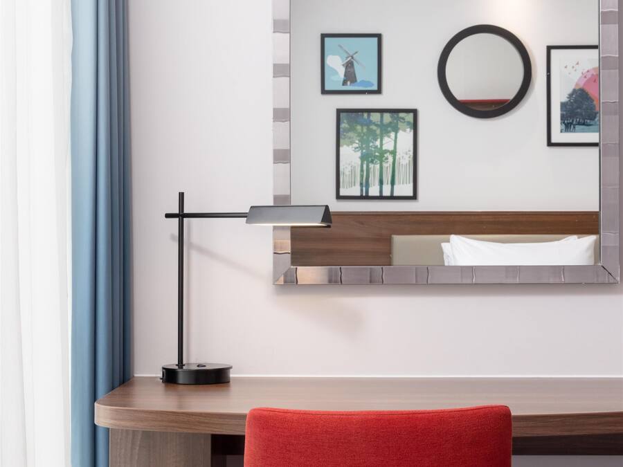 A hotel room with a wooden desk, a red chair, a wall-mounted mirror, and framed pictures.
