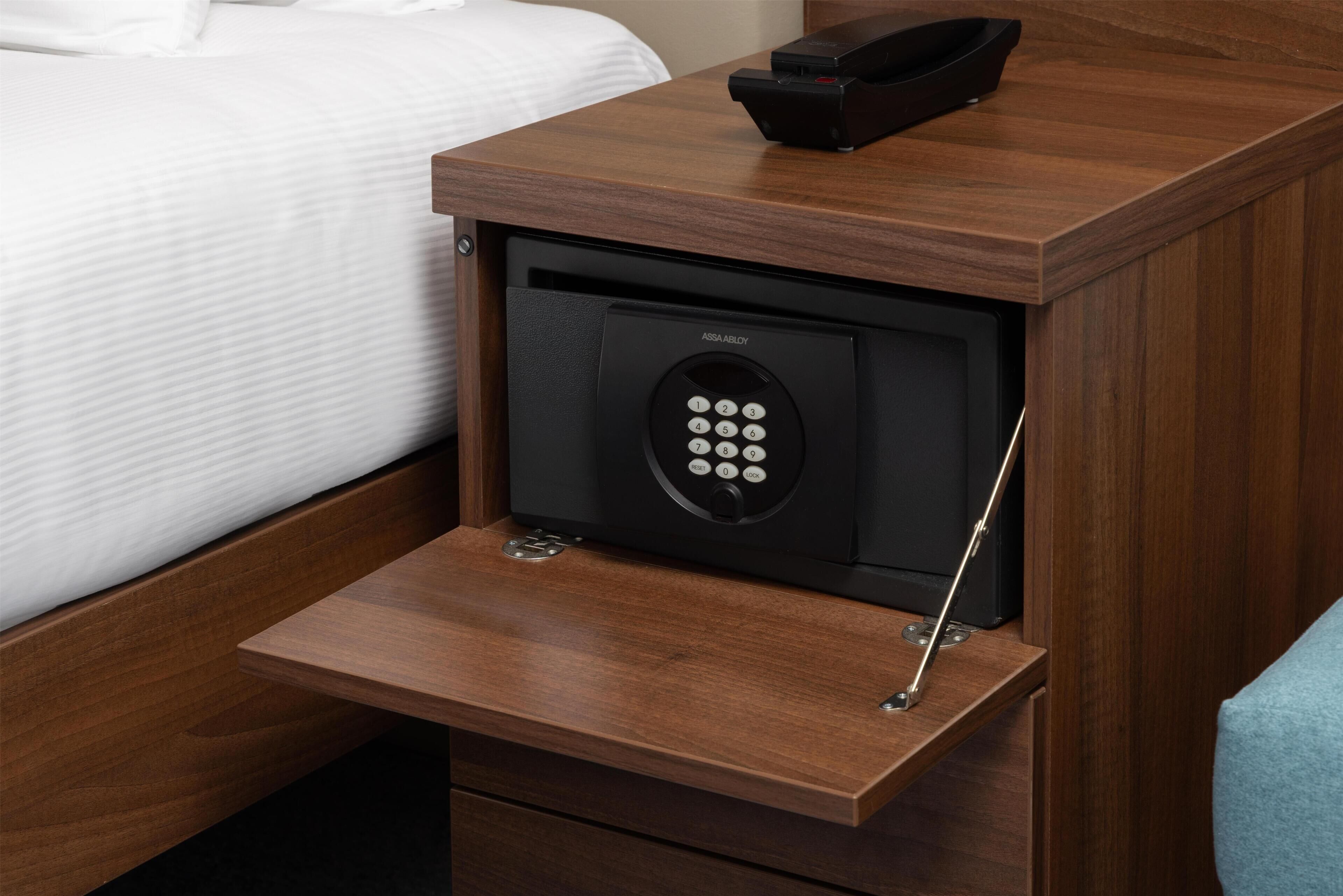 Habitación de hotel con mesita de noche de madera, caja fuerte empotrada y cama con ropa blanca.