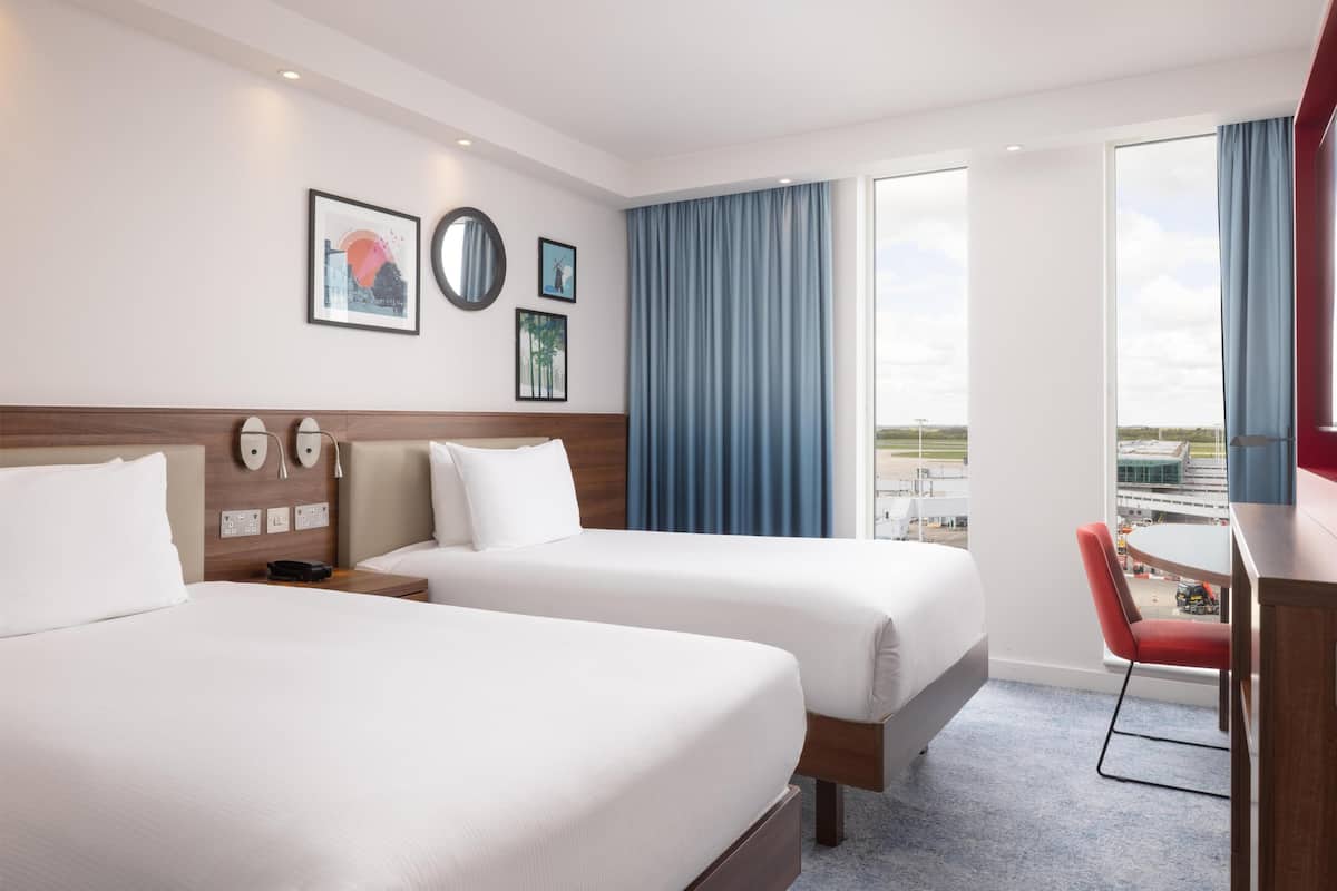 Habitación de hotel con dos camas, un escritorio con silla, un ventanal con cortinas y una obra de arte en la pared.