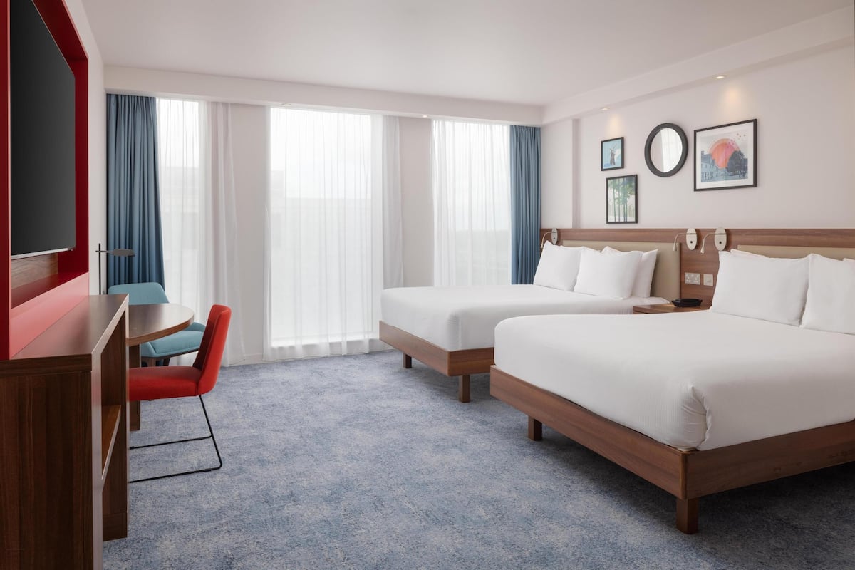 Habitación de hotel con dos camas, un escritorio, una silla y ventanas grandes con cortinas.
