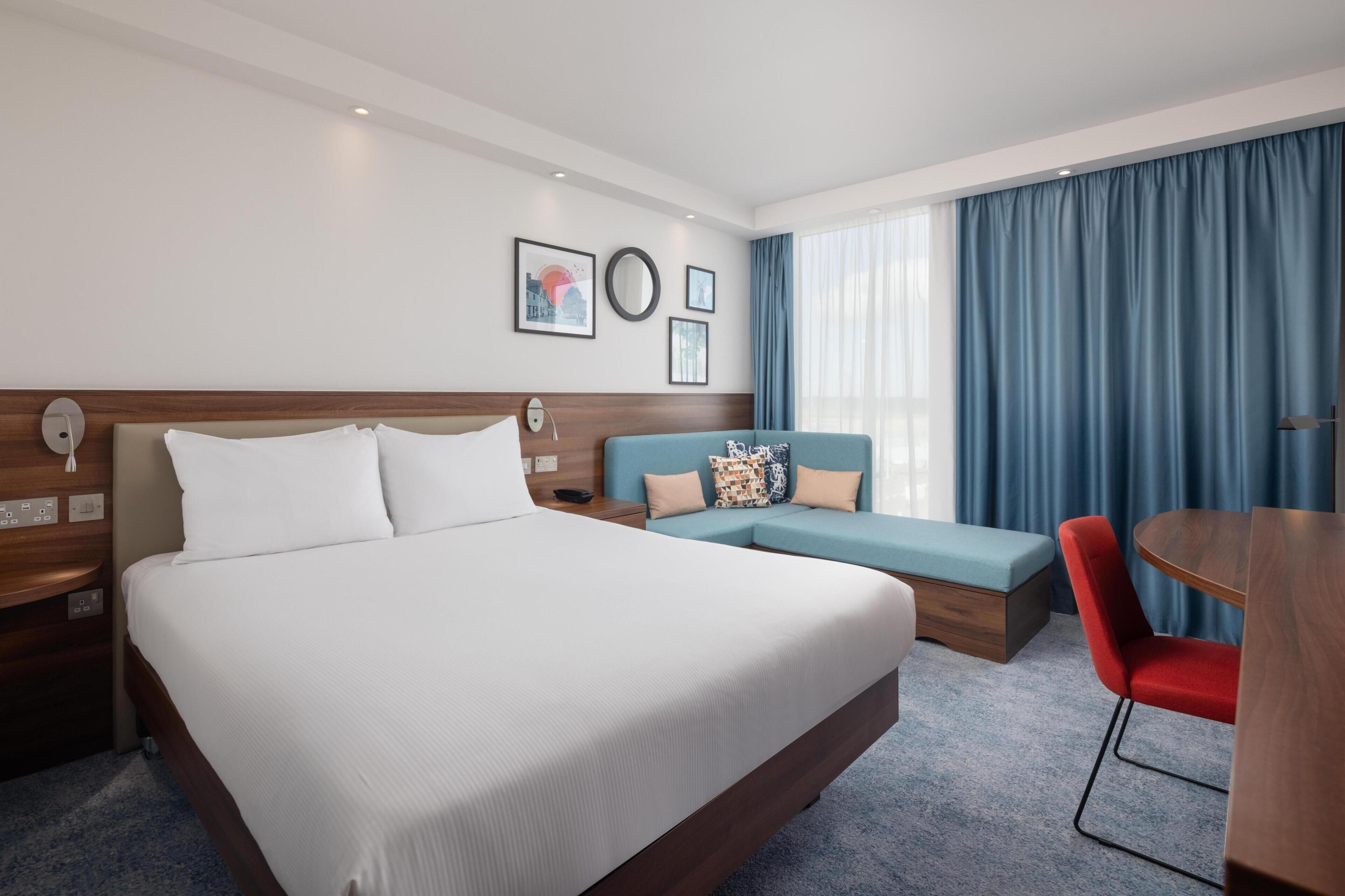 A hotel room with a large bed, a sofa, a red chair, a wooden table, and a blue curtain.