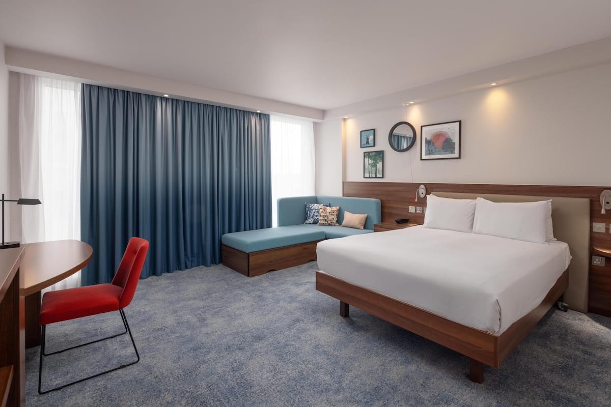 Habitación de hotel con cama, escritorio, una silla roja, un sofá azul y un ventanal con cortinas.
