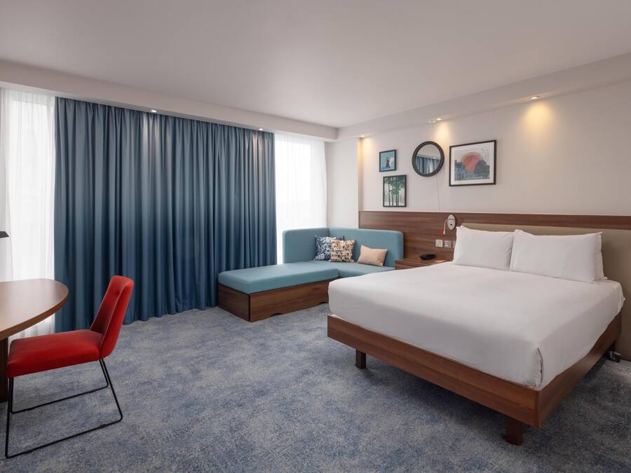A hotel room with a bed, a desk, a red chair, a blue sofa, and a large window with curtains.