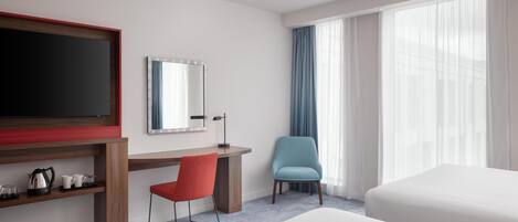 A hotel room with a bed, a desk with a chair, a TV, a mirror, and a window with curtains.