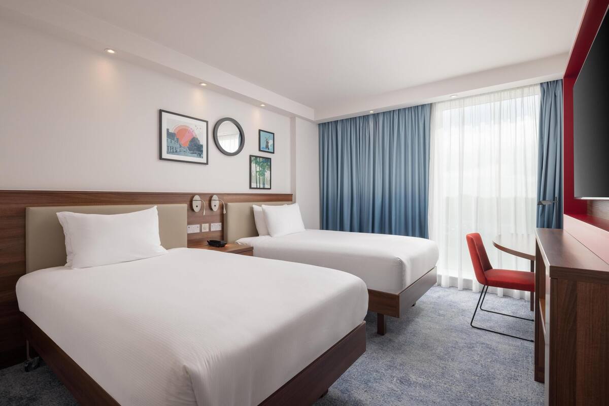Habitación de hotel con dos camas, un escritorio, una silla, un televisor y obras de arte en la pared.