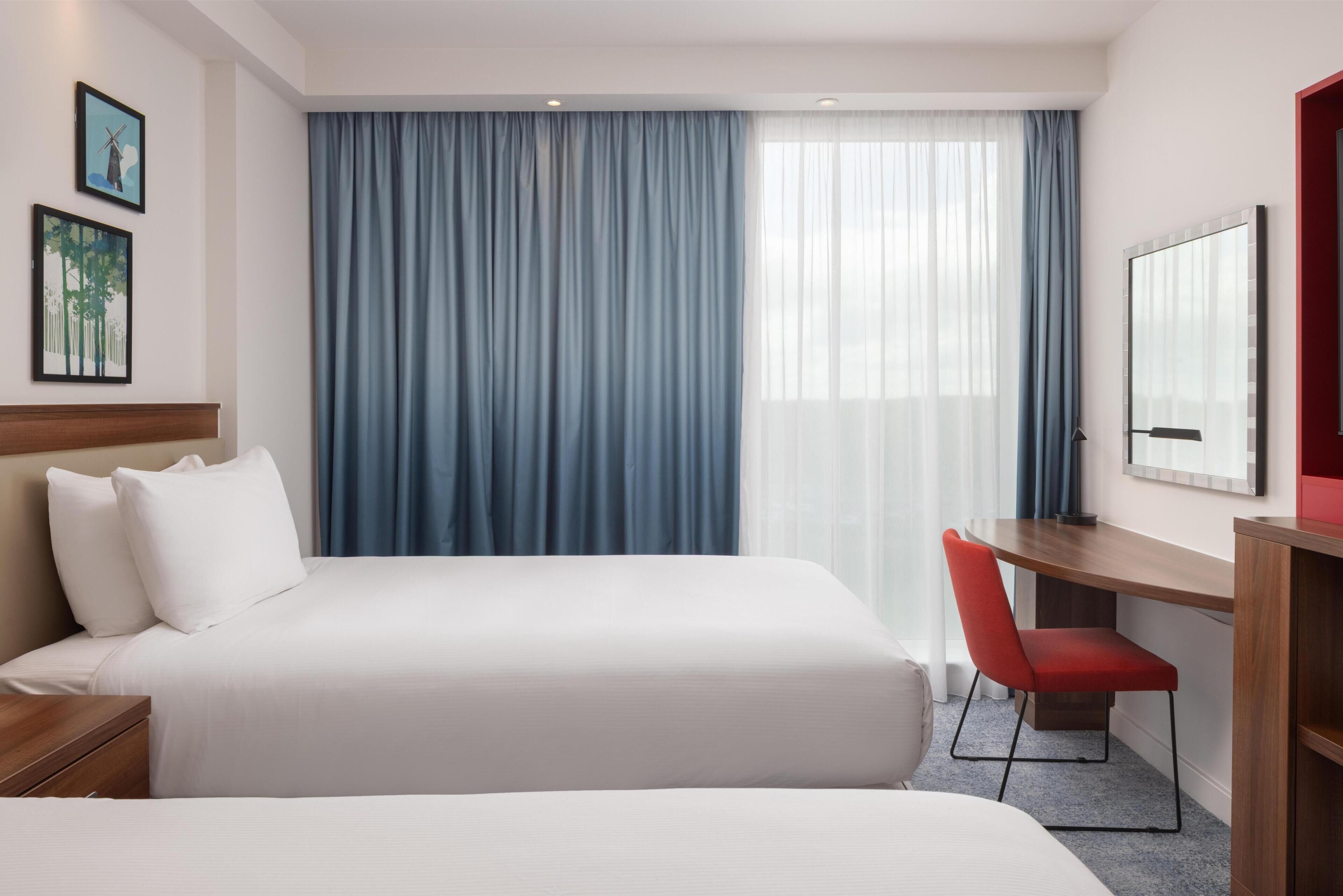A hotel room with two beds, a desk, a chair, a large window with curtains, and artwork on the walls.