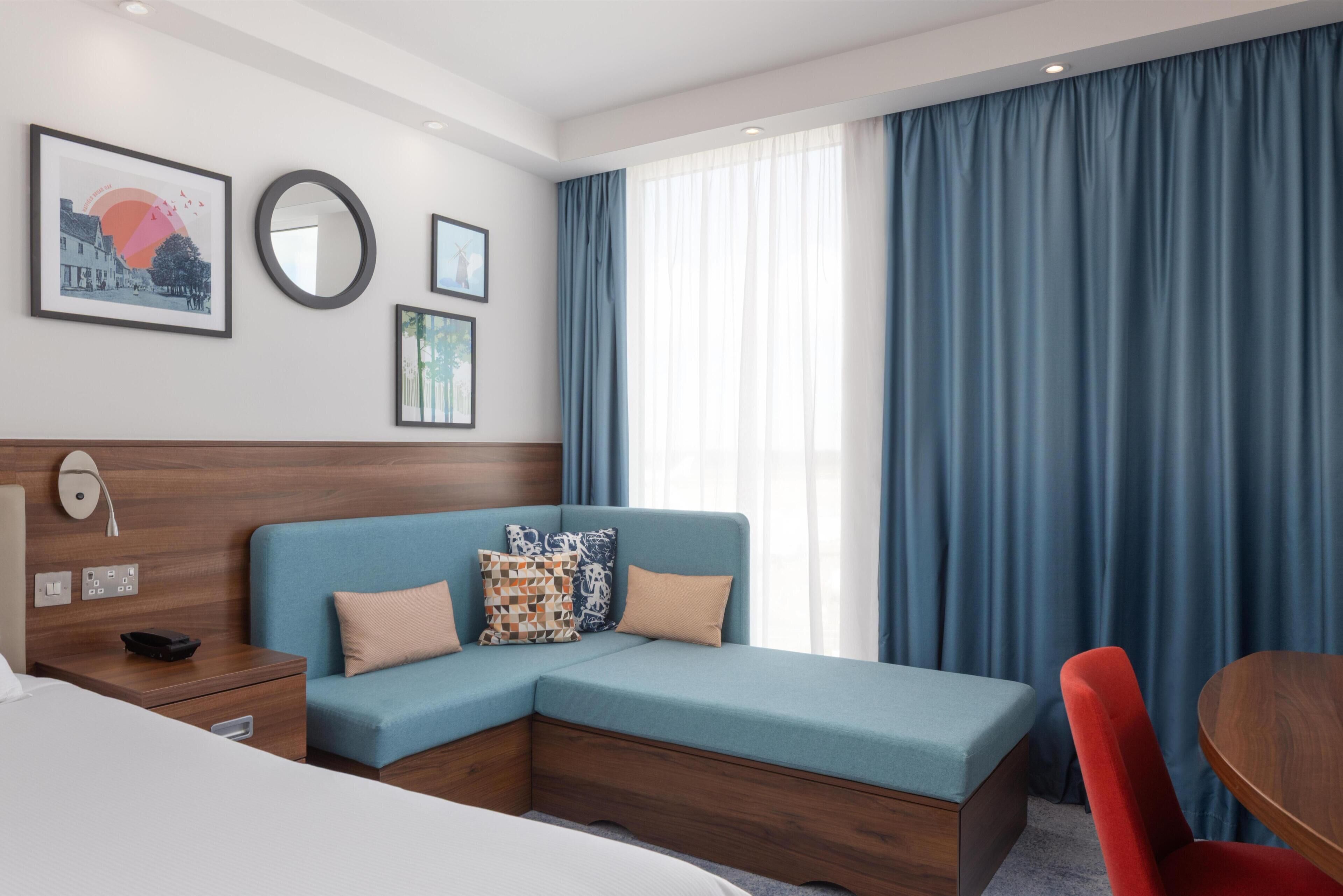 Una habitación de hotel moderna con un sofá modular, un cabecero de madera, una mesita de noche, una pequeña mesa redonda y un amplio ventanal con cortinas azules.