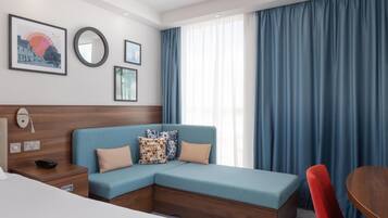 Una habitación de hotel moderna con un sofá modular, un cabecero de madera, una mesita de noche, una pequeña mesa redonda y un amplio ventanal con cortinas azules.