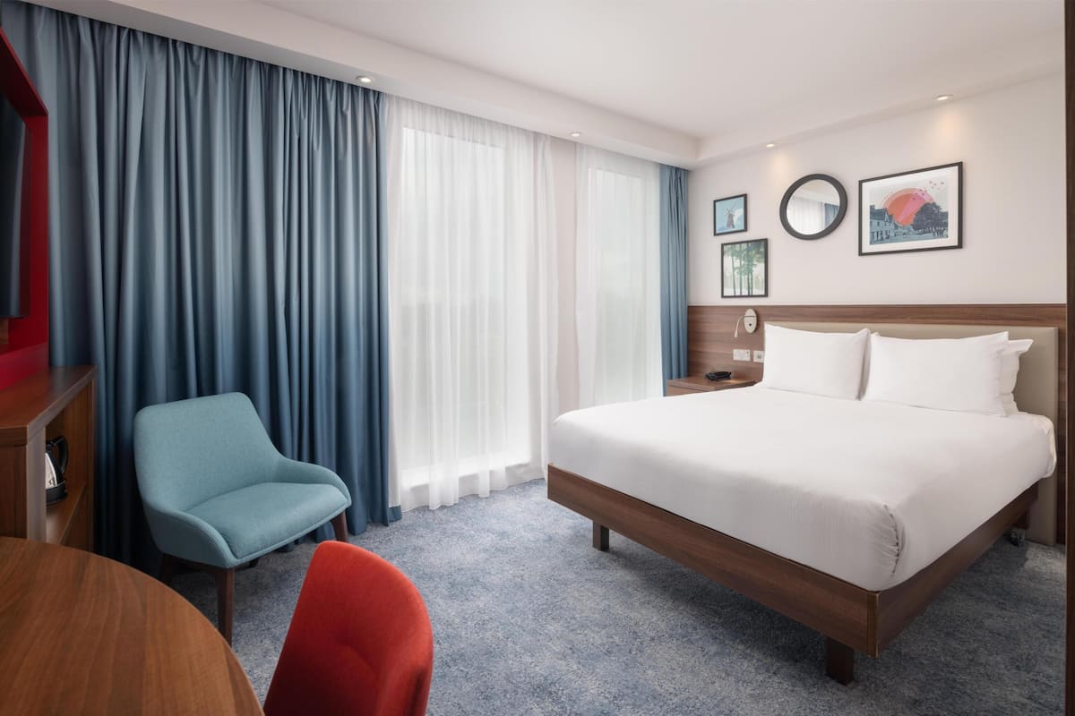 Habitación de hotel con cama, escritorio, silla y un ventanal con cortinas.
