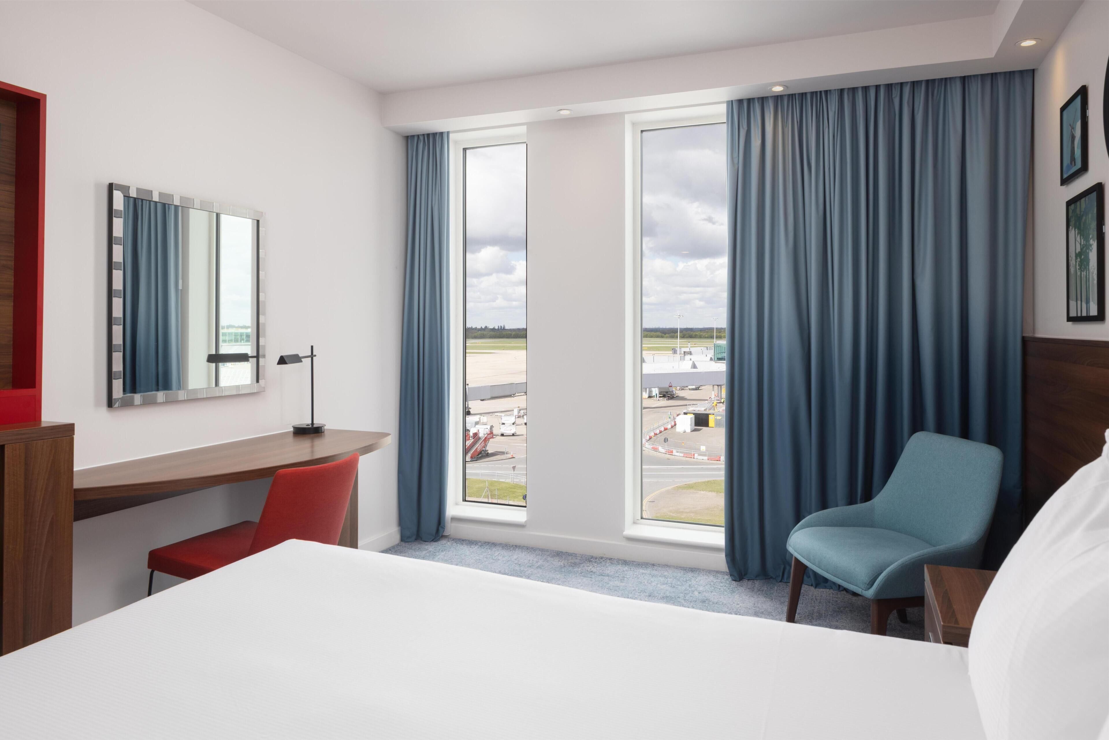 Habitació d'hotel amb un llit, una taula de treball, una cadira, un finestra gran amb cortines i vista a l'aeroport.