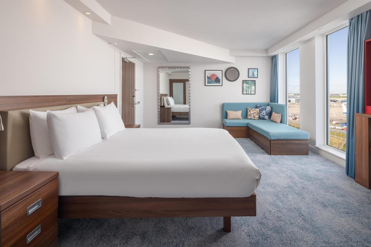 Habitación de hotel con una cama grande, un sofá, un escritorio y vista al aeropuerto.